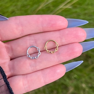 Könnte beinhalten: Zwei Nasenringe mit Kettengliedern, einer silberfarben und einer goldfarben, auf einer Hand gezeigt.