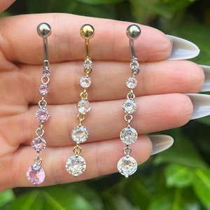Puede incluir: Tres piercings para el ombligo con colgantes. Uno es plateado con gemas rosas, otro es dorado con gemas transparentes y el tercero es plateado con gemas transparentes. Cada uno tiene una pequeña bola en la parte superior.