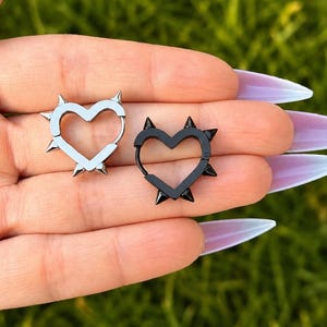Pode incluir: Dois brincos em forma de coração, um prateado e outro preto, cada um adornado com pequenas pontas. Os brincos são segurados em uma mão contra um fundo verde desfocado. O design de coração adiciona um toque ousado.