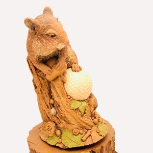 May include: A detailed figurine of a squirrel holding a golf ball, perched on a tree stump. The squirrel is brown with a textured finish, and the golf ball is white. The base of the stump has green leaves and the number 81.