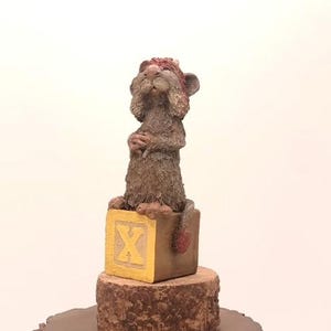 May include: A small, brown, furry animal figurine wearing a red hat sits on a yellow block with the letter "X" on it. The block is on a brown tree stump.