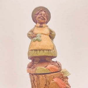 May include: A whimsical figurine of a woman wearing a green dress and a yellow apron, holding a four-leaf clover. She is standing on a tree stump with leaves and acorns.