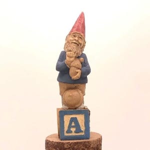 May include: A small gnome figurine with a red hat and blue jacket holding a small brown bear. The gnome is standing on a wooden block with the letter 'A' painted on it.