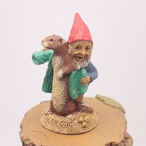 May include: A small gnome figurine wearing a red hat and blue jacket, holding a brown and white weasel figurine. The base of the figurine is brown and has the words "Mink Coat" written on it.