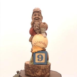 Könnte beinhalten: Eine Gnome-Figur, die eine Baseballkappe trägt und einen Baseball hält. Der Gnome sitzt auf einem Baseball und einem blauen Block mit der Nummer 9 darauf. Die Figur steht auf einem Holzsockel.