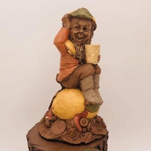 May include: A whimsical figurine of a gnome with a brown beard and a green hat, sitting on a yellow fruit with a brown base. The gnome is holding a brown cup.