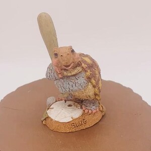 May include: A small ceramic figurine of a turtle holding a baseball bat. The turtle is wearing a blue and brown shell and has a brown head. The figurine is standing on a small brown base with the word "SLUG" written on it.
