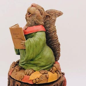 May include: A ceramic figurine of a squirrel wearing a green robe and a red scarf, holding a book titled "Carols for Critters". The squirrel is sitting on a tree stump base decorated with autumn leaves and acorns.
