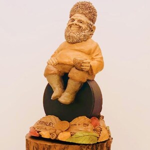 May include: A whimsical figurine of a gnome sitting on a black puck, holding a cup. The gnome is wearing a brown shirt and has a bushy beard. The base of the figurine is decorated with leaves and acorns. The figurine is labeled "Stanley".