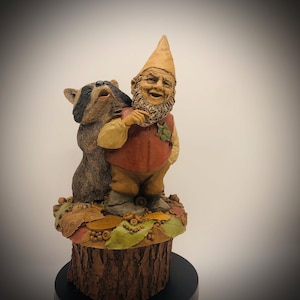 May include: A whimsical garden gnome figurine with a raccoon. The gnome wears a red waistcoat, tan hat, and has a long white beard. The raccoon is brown and black. The base is a tree stump with leaves and acorns.