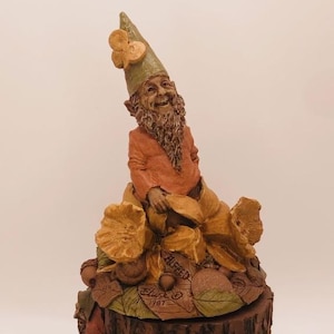 May include: A whimsical gnome figurine with a red and orange outfit, sitting on a tree stump with a mushroom and leaf design. The gnome is wearing a green hat with a butterfly design and has a long white beard. The base of the figurine is inscribed with "Black & Co. 1987".