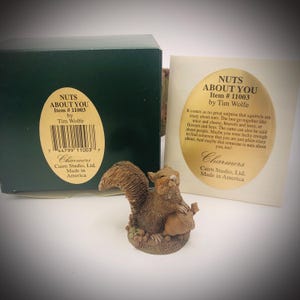 May include: A brown squirrel figurine holding a nut, with the base inscribed with the text NUTS ABOUT YOU. Behind it sits a dark green box and a white card, both featuring an oval gold label with the text NUTS ABOUT YOU Item # 11003 by Tim Wolfe.
