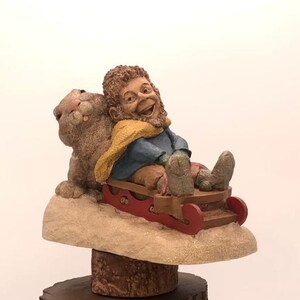 Puede incluir: Una figurita caprichosa de un gnomo con barba marrón y una bufanda amarilla montando un trineo rojo con un conejo marrón. La figurita está sentada sobre una base de madera marrón.
