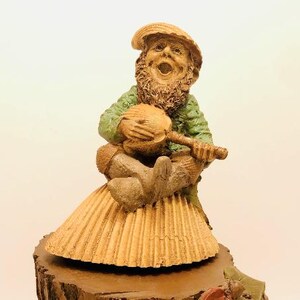 May include: A whimsical figurine of a gnome with a brown beard and a green shirt, holding a brown musical instrument. The gnome is sitting on a brown base with a textured, layered skirt.