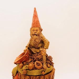 May include: A whimsical gnome figurine with a red hat sits on a tree stump surrounded by acorns. The gnome is holding a small bucket and has a friendly expression.