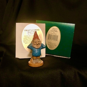 May include: A small, blue and red gnome figurine with a white beard and a red hat. The gnome is holding its hands over its ears. The figurine is on a base that says "That's Corny". The figurine is part of a collection called "Charmers" by Cairn Studio, Ltd. and is made in America.