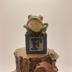 May include: A green frog figurine perched atop a blue block with the letter "F". The frog is sitting on a brown tree stump with a leaf and a small acorn. The figurine has a whimsical, decorative design.