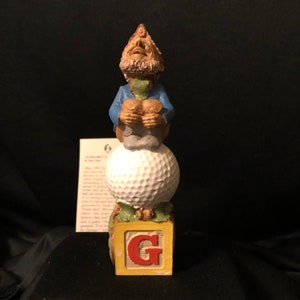 May include: A small ceramic figurine of a gnome sitting on top of a golf ball. The gnome is wearing a blue shirt and has a white beard. The golf ball is sitting on top of a wooden block with the letter "G" on it. The block is painted yellow with red letters.