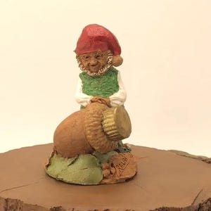May include: A small gnome figurine with a red hat and green vest is holding a large acorn. The gnome is standing on a brown base with the word "REPLICA" written on it.