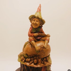 May include: A whimsical gnome figurine with a red and green hat sits on a brown mushroom with the text "Spud & Co." on the side. The gnome is holding a small leaf in its hand and has a smiling face.