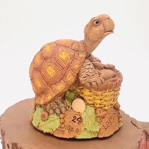 Op de afbeelding: Een klein, bruin en geel keramiek schildpadbeeldje dat op een bruine basis zit met groene bladeren en eikels. De schildpad houdt een klein mandje vast met een bruin en geel geweven patroon. De basis heeft de tekst "Wolfe 1993" en "Love" erop.