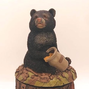 Puede incluir: Una figura de oso negro sosteniendo un tarro de miel. El oso está sentado sobre una base de tocón de árbol con hojas y bellotas. La figura tiene una apariencia detallada y texturizada y está pintada con colores naturales.