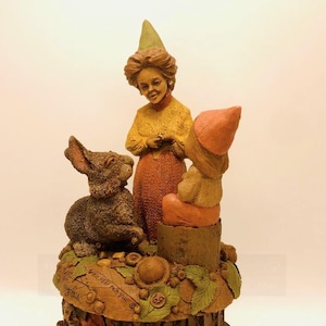 May include: A ceramic figurine set featuring a brown rabbit, a girl wearing a green and yellow hat, and a girl wearing a pink hat. The figurines are sitting on a tree stump base with leaves and acorns.