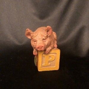 May include: A small, detailed figurine of a light brown pig resting on a yellow alphabet block with the letter "P". The pig has a pink snout and ears. The block has a light green leaf on the side.