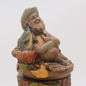 May include: A ceramic figurine of a gnome with a green hat, holding a fish and sitting on a bed of leaves and acorns. The gnome is smiling and has a long white beard. The base of the figurine is a tree stump.