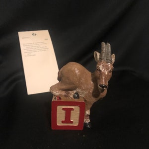 Puede incluir: Una figurita de cabra montés marrón y blanca de pie sobre un bloque rojo y crema con la letra "I" en él.