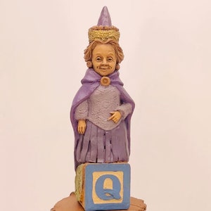 Puede incluir: Una figurita de una reina vestida de púrpura, con una corona dorada y un sombrero puntiagudo, de pie sobre un bloque azul con la letra "Q" en él.