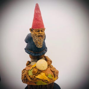 May include: A whimsical garden gnome statue with a red pointed hat, blue jacket, and long, tan beard. The gnome stands atop a detailed base featuring a golf ball, leaves, and acorns. The statue is made of a durable material.