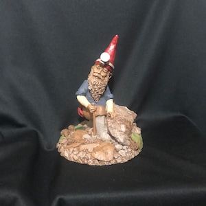 May include: A gnome figurine wearing a red hat and blue shirt is holding a pickaxe and standing on a rock formation with mushrooms.