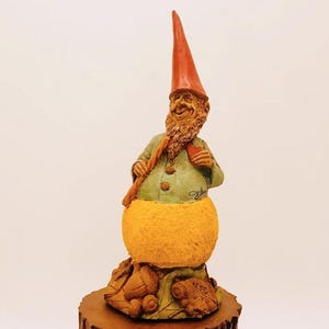 May include: A whimsical gnome figurine with a red hat, green shirt, and a yellow orange body. The gnome is holding a stick and is standing on a wooden base with acorns and leaves.