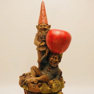 May include: A whimsical figurine featuring two garden gnomes. One gnome stands, wearing a red pointed hat and a brown coat, while the other reclines, supporting a large red apple. The base is adorned with leaves.