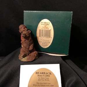 May include: A small figurine of a gnome hugging a brown bear. The figurine is made of painted resin and is part of the Charmers collection. The text on the box reads "Bearback Item # 12002 by Tom Clark and Tim Wolfe".