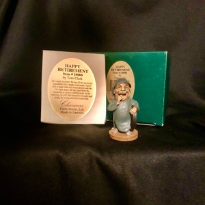 May include: A figurine of a man with a moustache and a surprised expression, wearing a blue shirt and holding a tyre. The figurine is made of painted resin and is sitting on a wooden base. The figurine is packaged in a green box with the words "Happy Retirement" and "Item # 10008" printed on it.