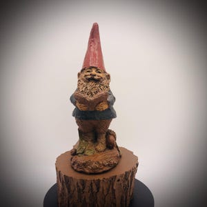 May include: A garden gnome statue with a red pointed hat, holding a book. The gnome has a long, brown beard and is wearing a blue jacket and brown pants. The statue is standing on a brown, tree-stump base.