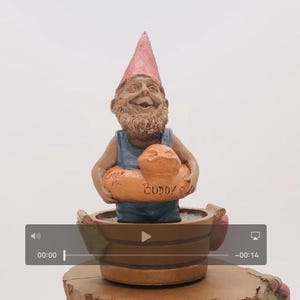 May include: A gnome figurine wearing a red hat and blue swimsuit holds a bright orange inner tube with the text "Buddy O" on it. The gnome is sitting in a brown wooden bucket.