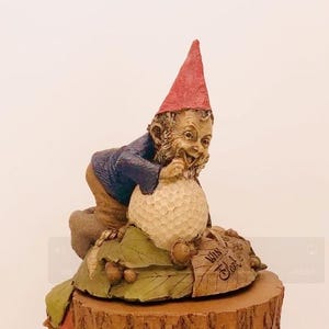 May include: A whimsical gnome figurine with a red hat, blue shirt, and a mischievous grin, holding a golf ball. The gnome is sitting on a tree stump with leaves and acorns. The text "Why Elfin Golf" is written on a sign.