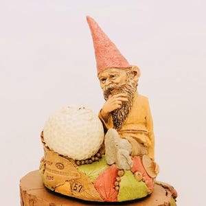 May include: A whimsical gnome figurine with a pink hat, a white beard, and a brown robe, sitting next to a white golf ball. The gnome is holding his chin with his hand and looking at the golf ball. The figurine is surrounded by colorful leaves and a number "57" on the base.