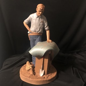 May include: A detailed figurine of a man in a light blue shirt and blue pants, leaning on a work table. The figurine has a brown base with tools and a shovel. The man is looking down at the work table.