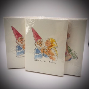 May include: A set of three white greeting cards wrapped in clear plastic. The center card features a watercolor illustration of a gnome with a red hat and a person with a yellow headscarf, with the text THANK YOU printed below the figures.