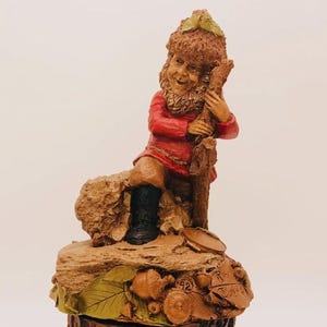 May include: A whimsical figurine of a gnome, dressed in a red jacket and black boots, leaning against a wooden post. The gnome has a detailed face and is perched on a base with leaves and acorns. The figurine is a charming decorative item.