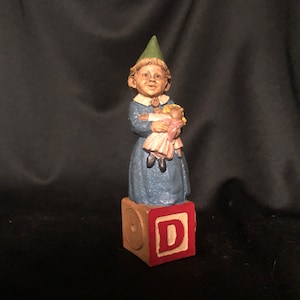 May include: A ceramic figurine of a girl gnome wearing a green hat and a blue dress, holding a doll. The gnome is standing on a wooden block with the letter 'D' painted on it.