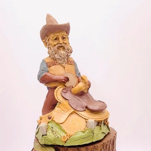 May include: A figurine of a gnome wearing a brown cowboy hat and a vest, sitting on a saddle with a brown leather seat and a yellow and brown base. The base has the word "Two Bits" written on it.