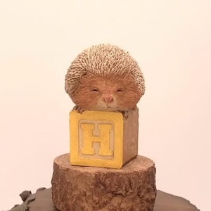 May include: A brown and white hedgehog figurine with closed eyes is sitting on top of a yellow block with the letter "H" on it. The block is sitting on a brown tree stump.