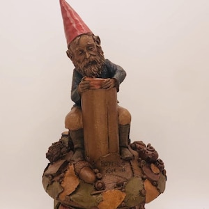 May include: A whimsical gnome figurine with a red hat sits atop a mushroom-shaped base. The gnome is holding a small pot and is surrounded by various woodland elements, including leaves, acorns, and a teapot. The base is decorated with a signature that reads "Potter ©  Bark 372".