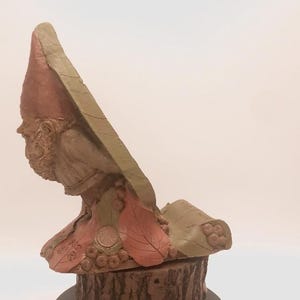 May include: A terracotta sculpture of a person wearing a pointed hat and a cloak. The sculpture is detailed with leaves and berries and is sitting on a wooden base.