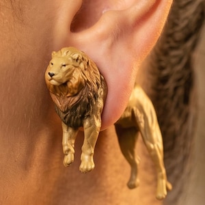 May include: Gold-colored lion earrings. The earrings are designed to appear as if the lion is walking through the ear. The lion's mane is a darker brown color. The earrings are a unique and eye-catching accessory.
